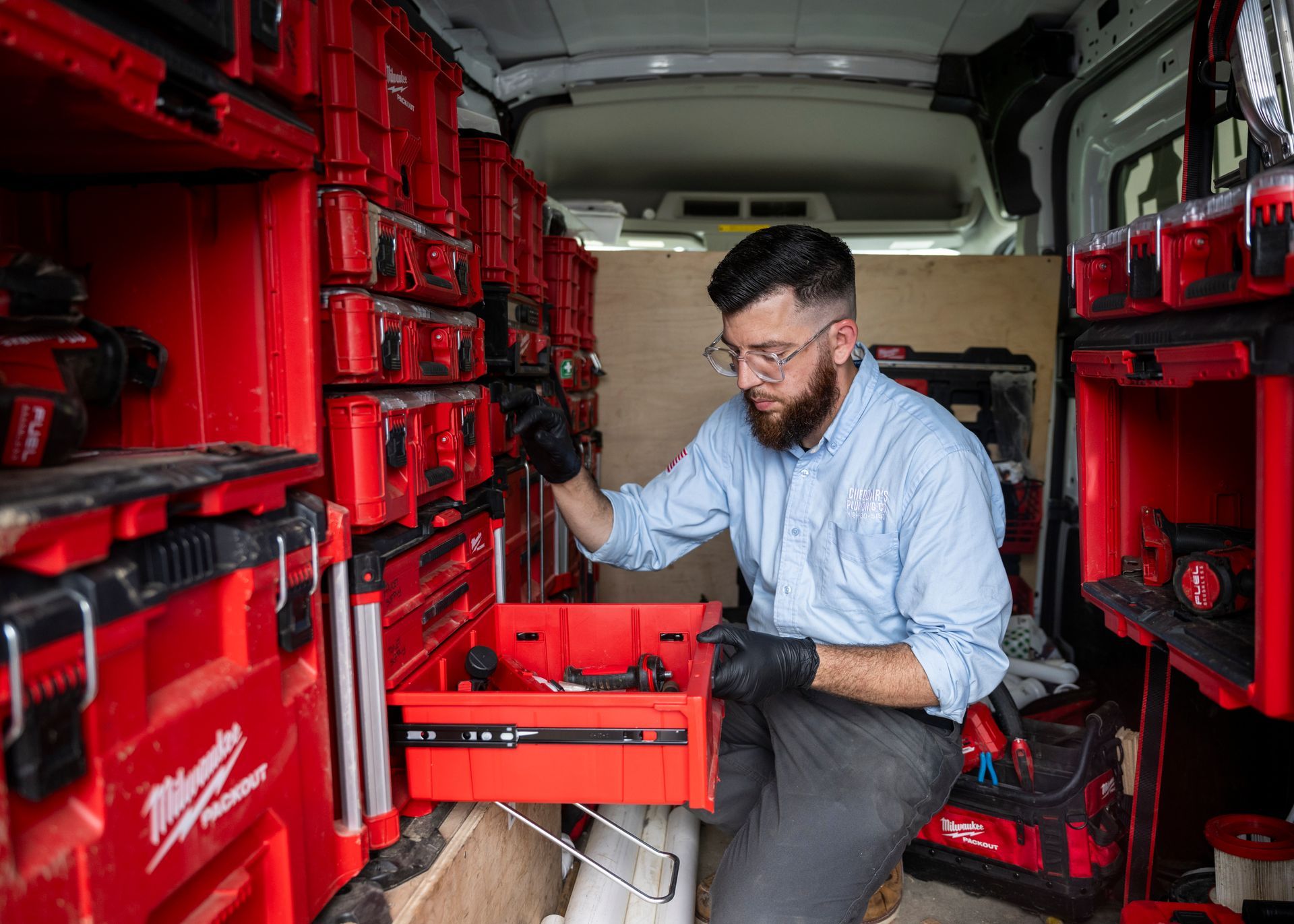 A person wearing gloves and glasses opens a red toolbox in a van filled with organized red toolboxes.