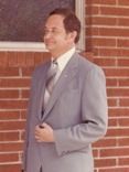 Man in a gray suit stands in front of a brick wall, holding his jacket lapel, smiling.