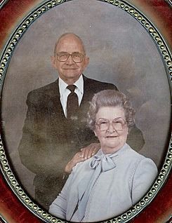 Elderly couple portrait in decorative frame; man in suit, woman in light blouse.