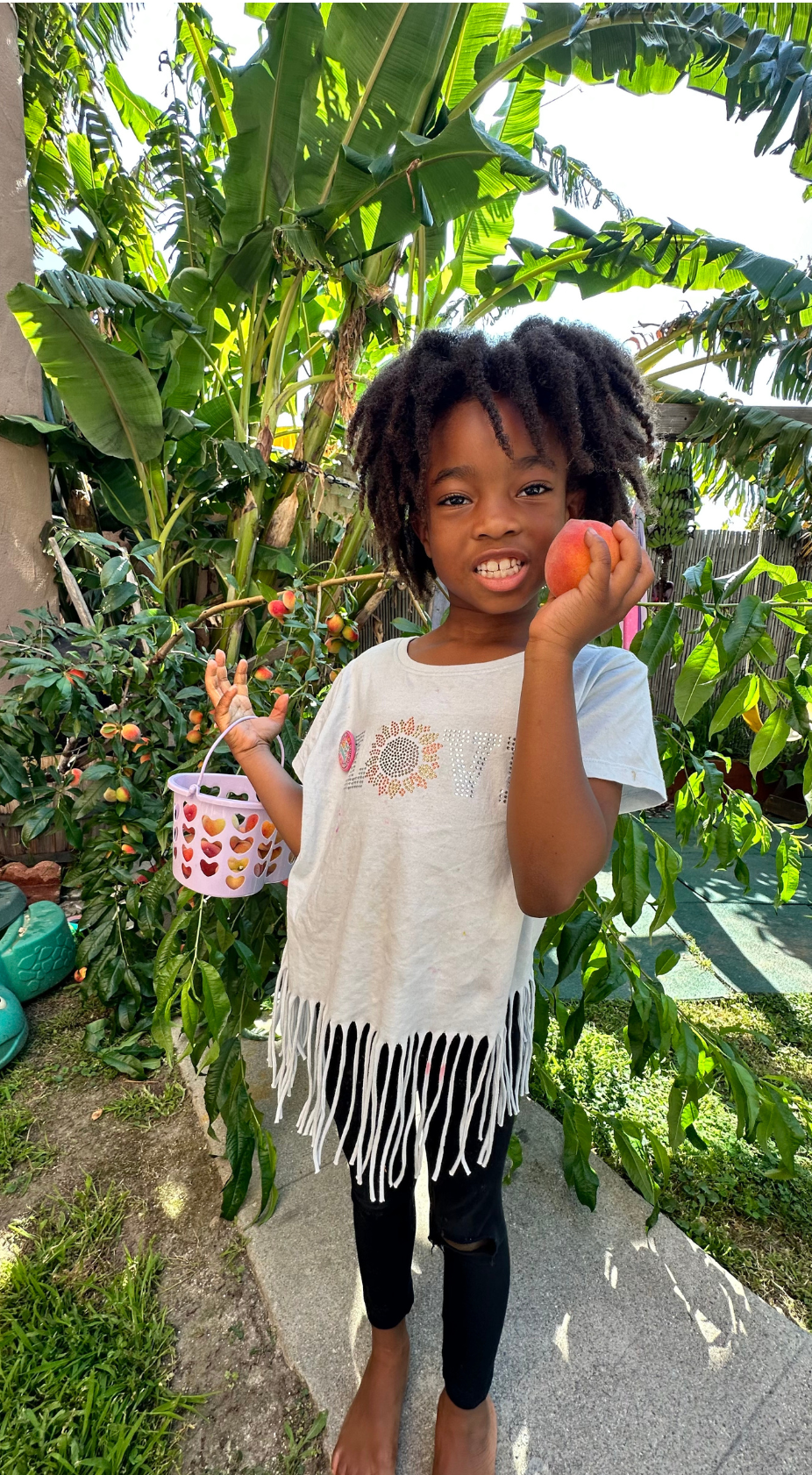 A little girl is holding a peach in her hand in front of a tree. Eden Creative Arts - Private Christian Micro School 