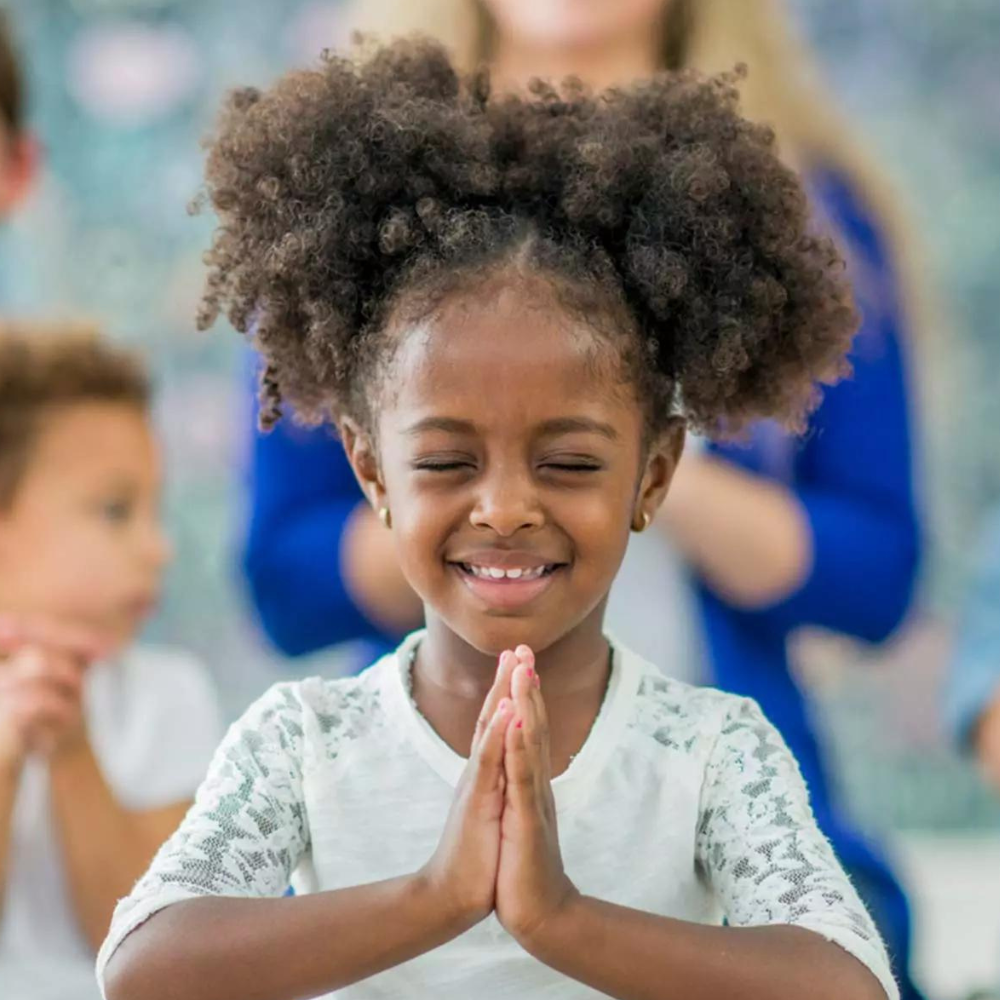 A little girl is smiling with her eyes closed and her hands folded in prayer. Eden Creative Arts