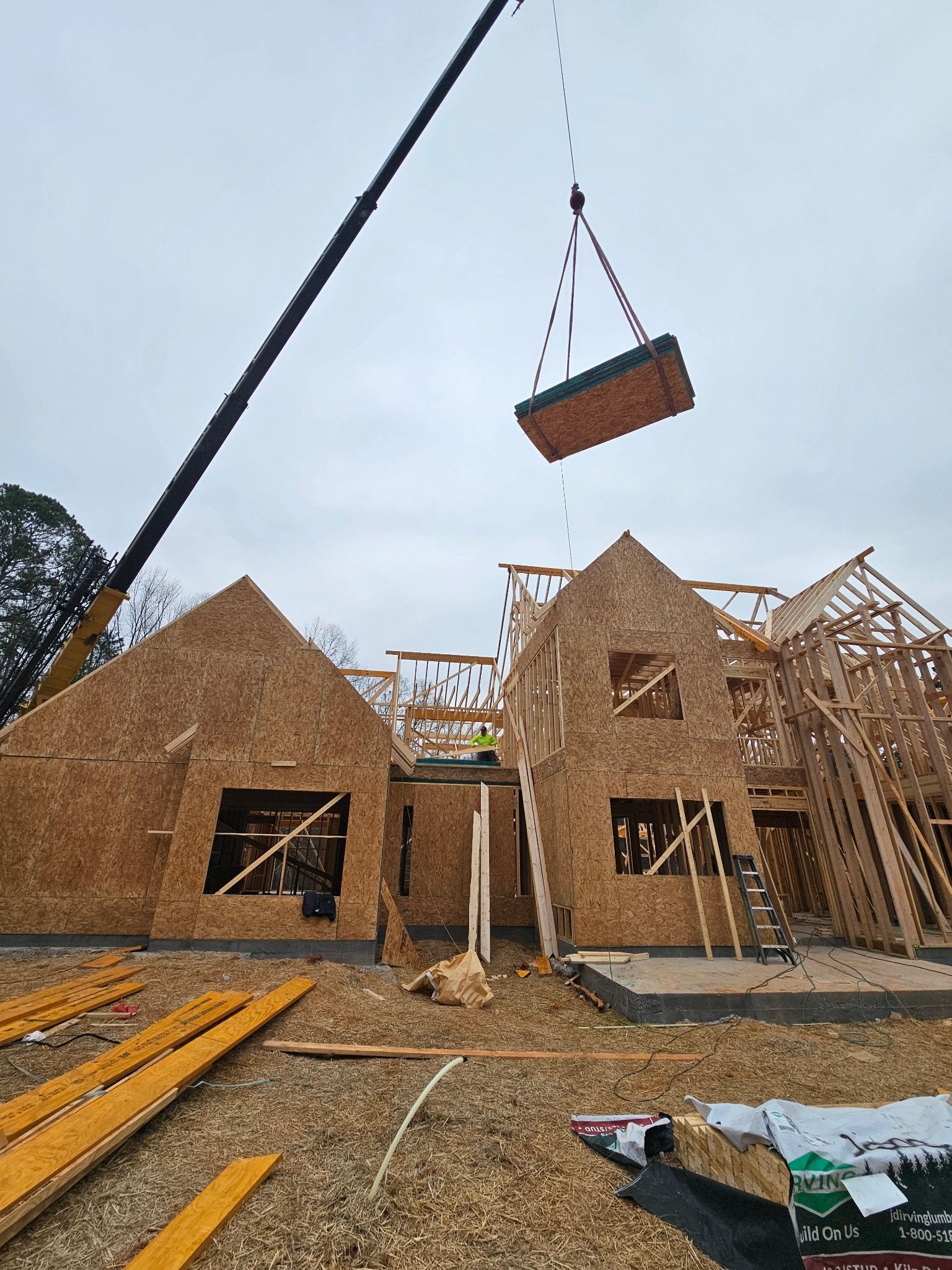 A crane is lifting a large piece of wood in front of a house under construction.