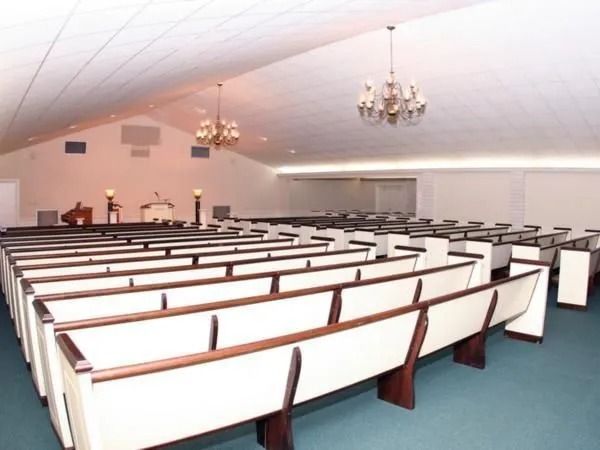 Empty church sanctuary with rows of white pews, chandeliers, and a raised platform.