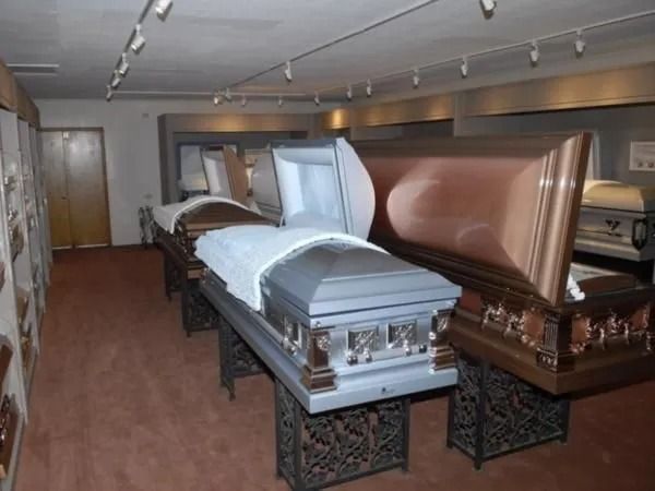 Coffin showroom with several open caskets on display, carpeted floor, overhead lighting.