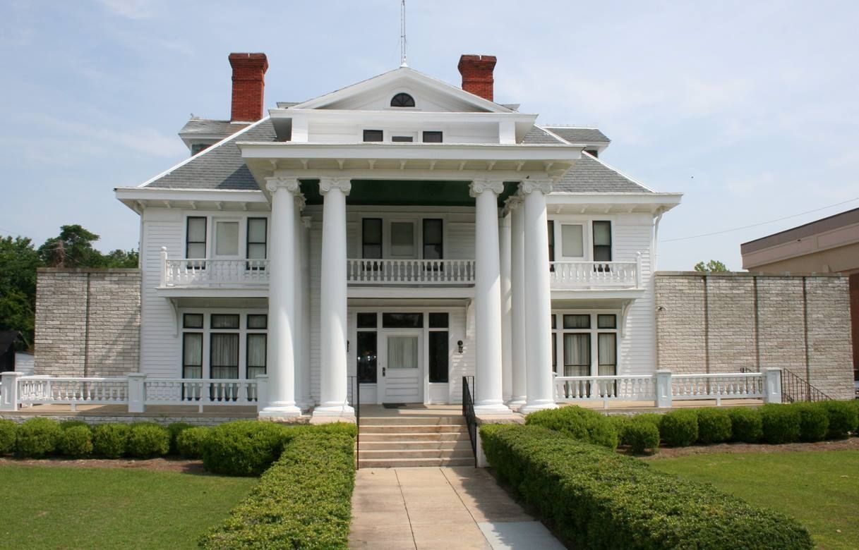 White two-story house with large columns on the porch, green lawn, bushes, and a pathway leading to the front door.