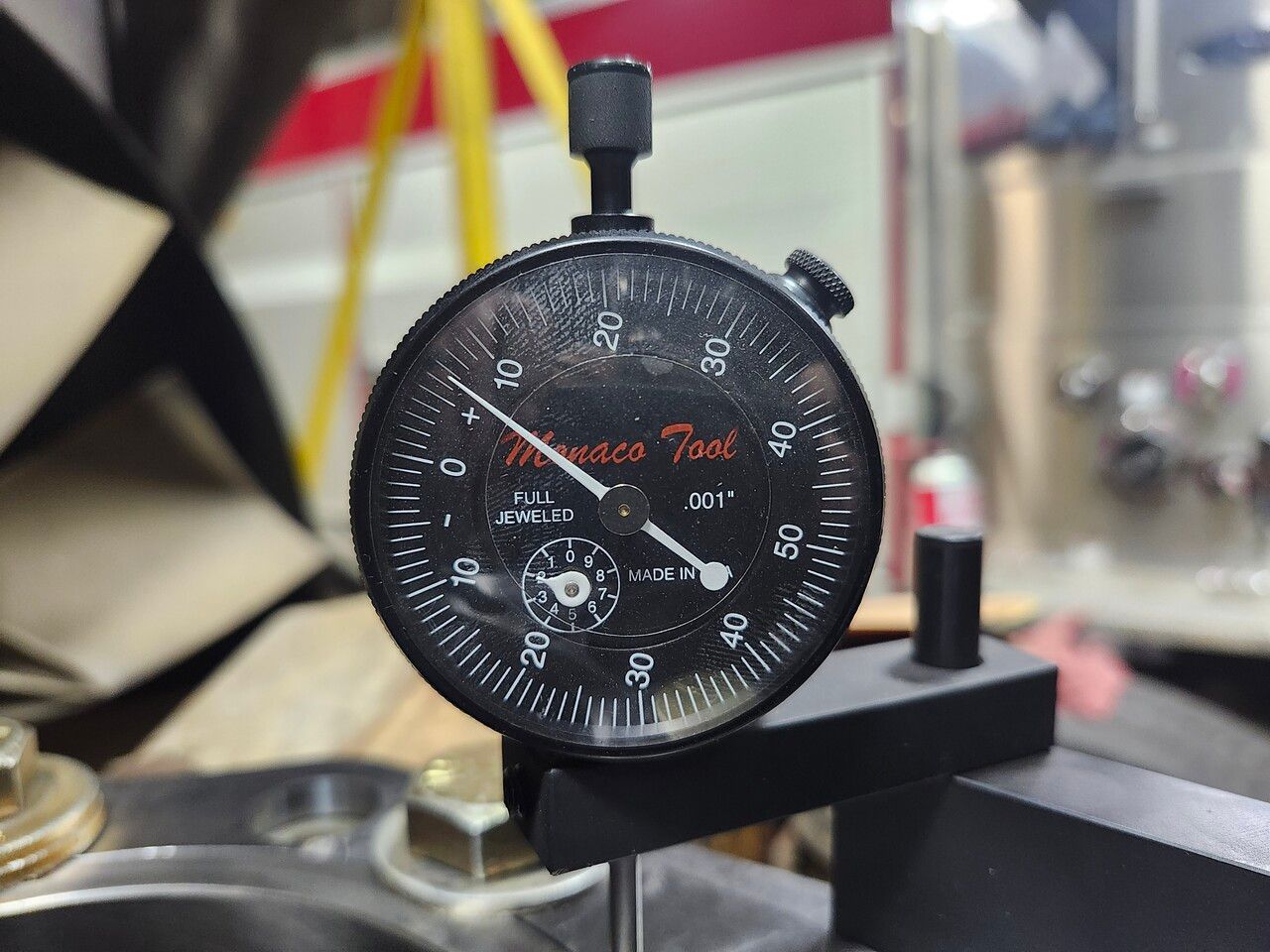 A black Monaco Tools dial indicator gauge mounted on a stand, showing a measurement with the needle pointing near 7.