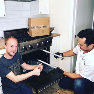 Two men installing oven racks in a kitchen; one gives a thumbs-up, the other holds a rack; cardboard box on the stovetop.