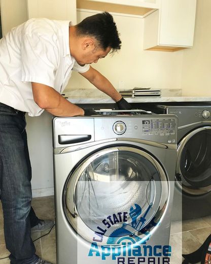 A repairman is working on a silver washing machine in a laundry room. He wears a white shirt and jeans.