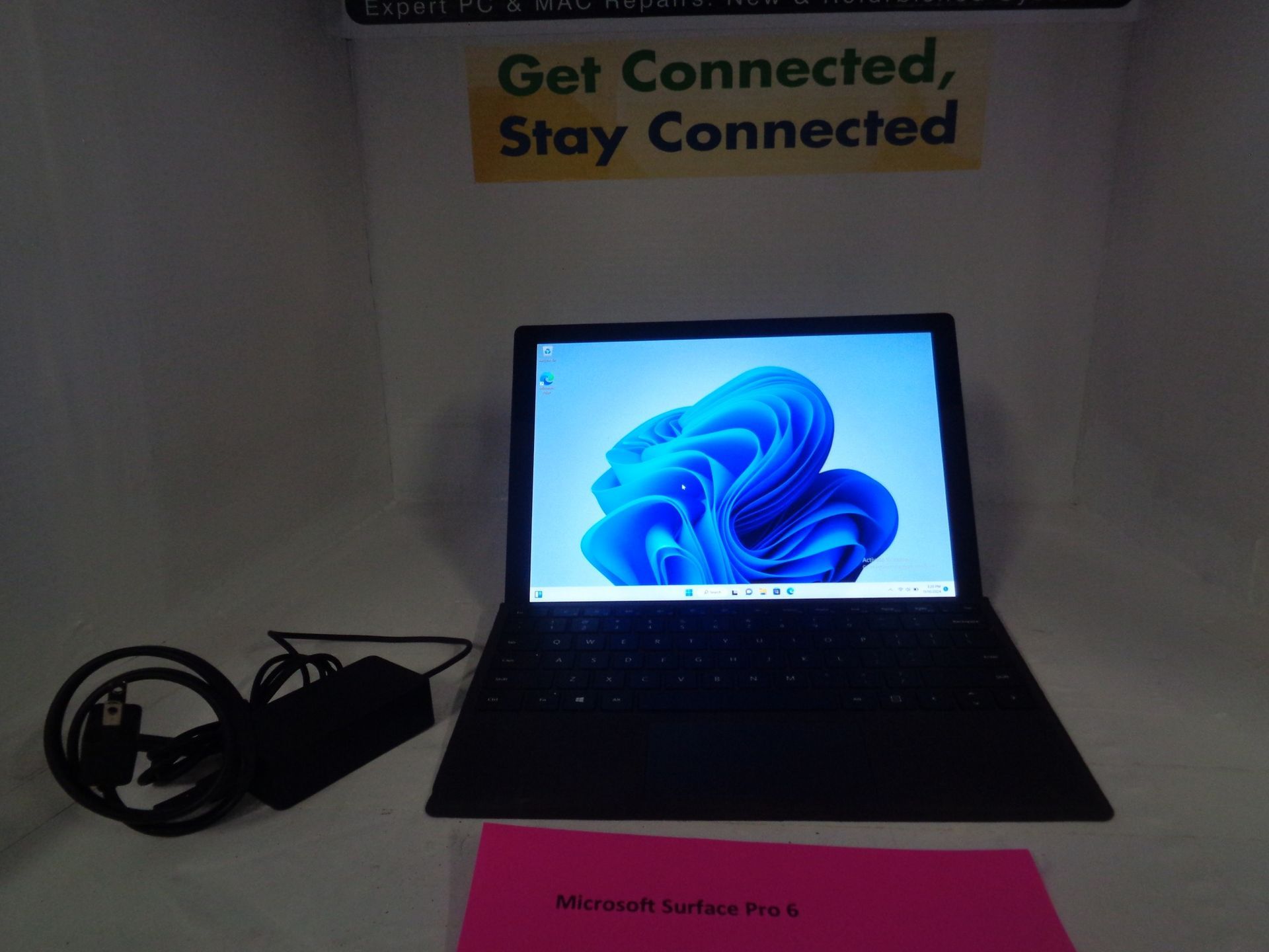 Microsoft Surface Laptop on a table with power adapter and pink pamphlet; blue Windows 11 wallpaper.