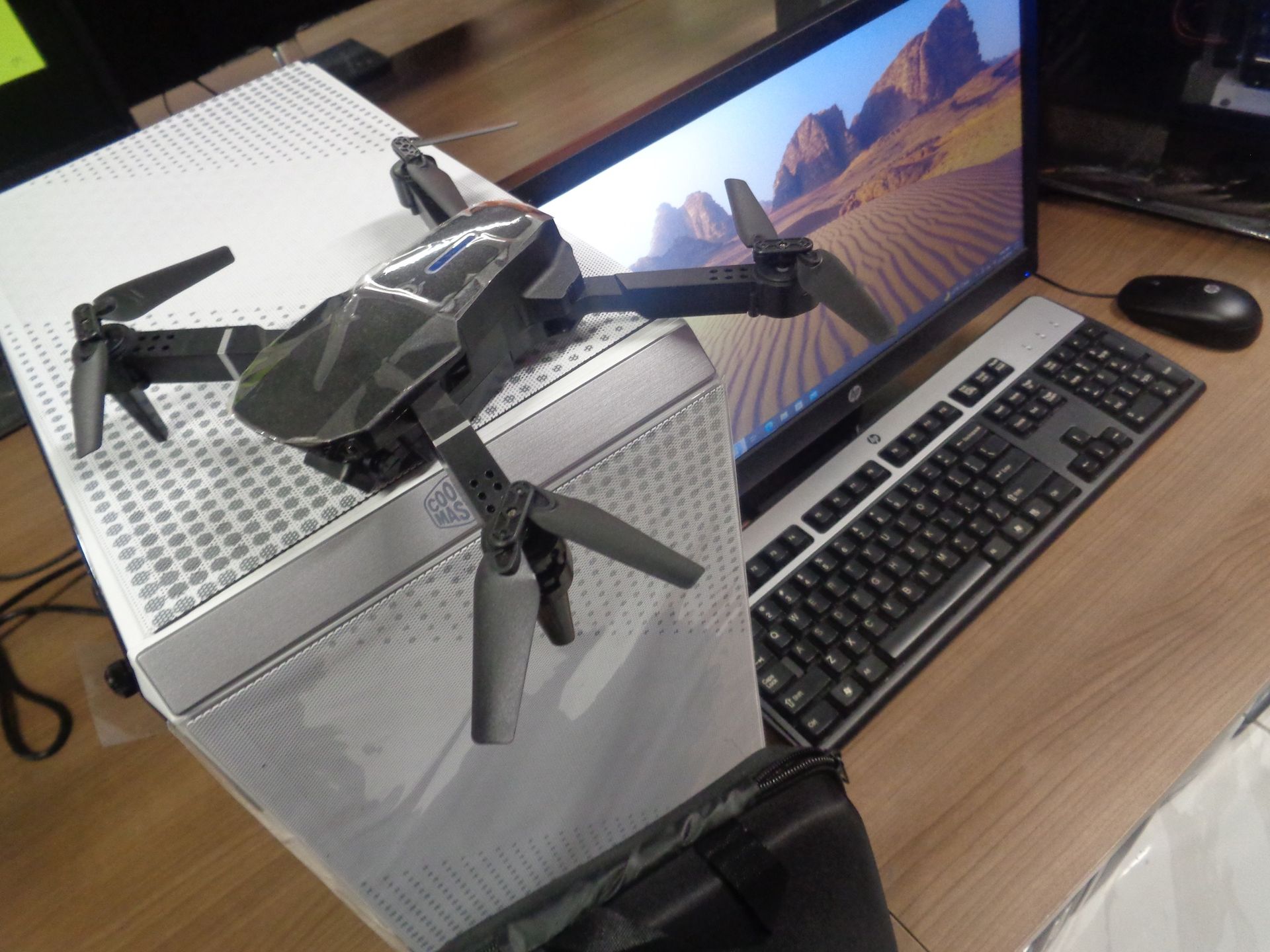 Black drone resting on a white computer tower next to a monitor, keyboard, and mouse on a wooden desk.
