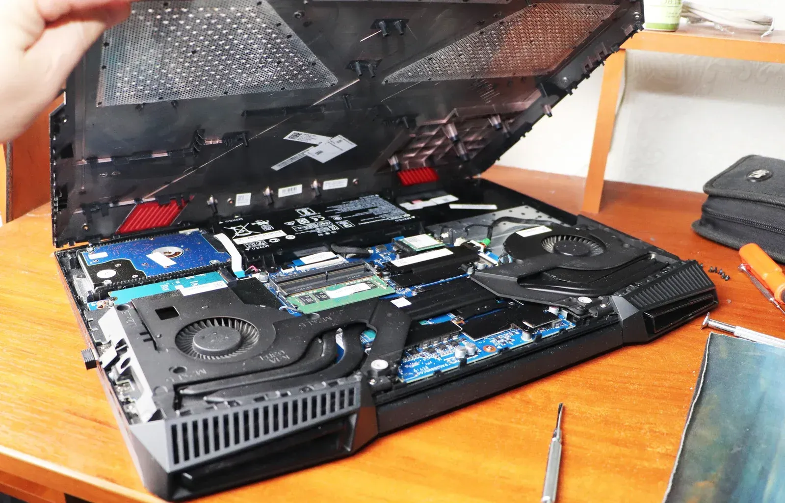 Laptop's top cover lifted, revealing internal components: motherboard, fans, and RAM. Someone's hand is visible.