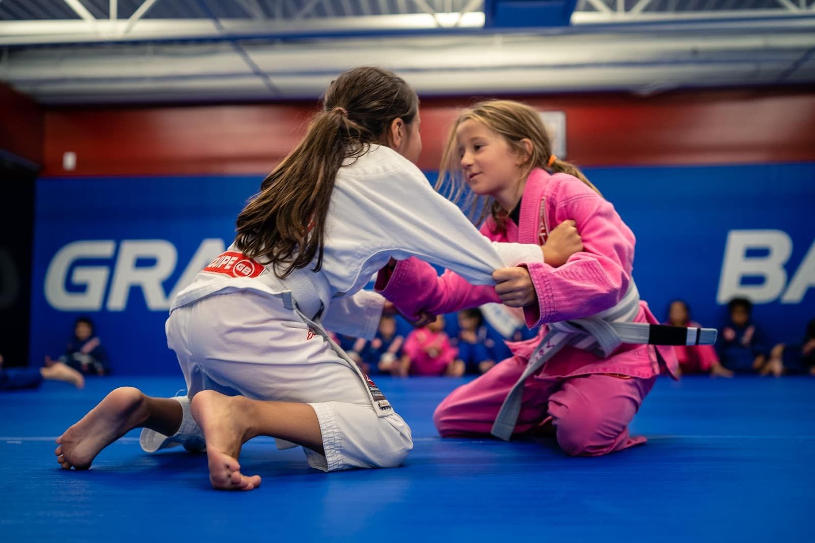 Two young girls are practicing jiu jitsu on a blue mat in a gym.