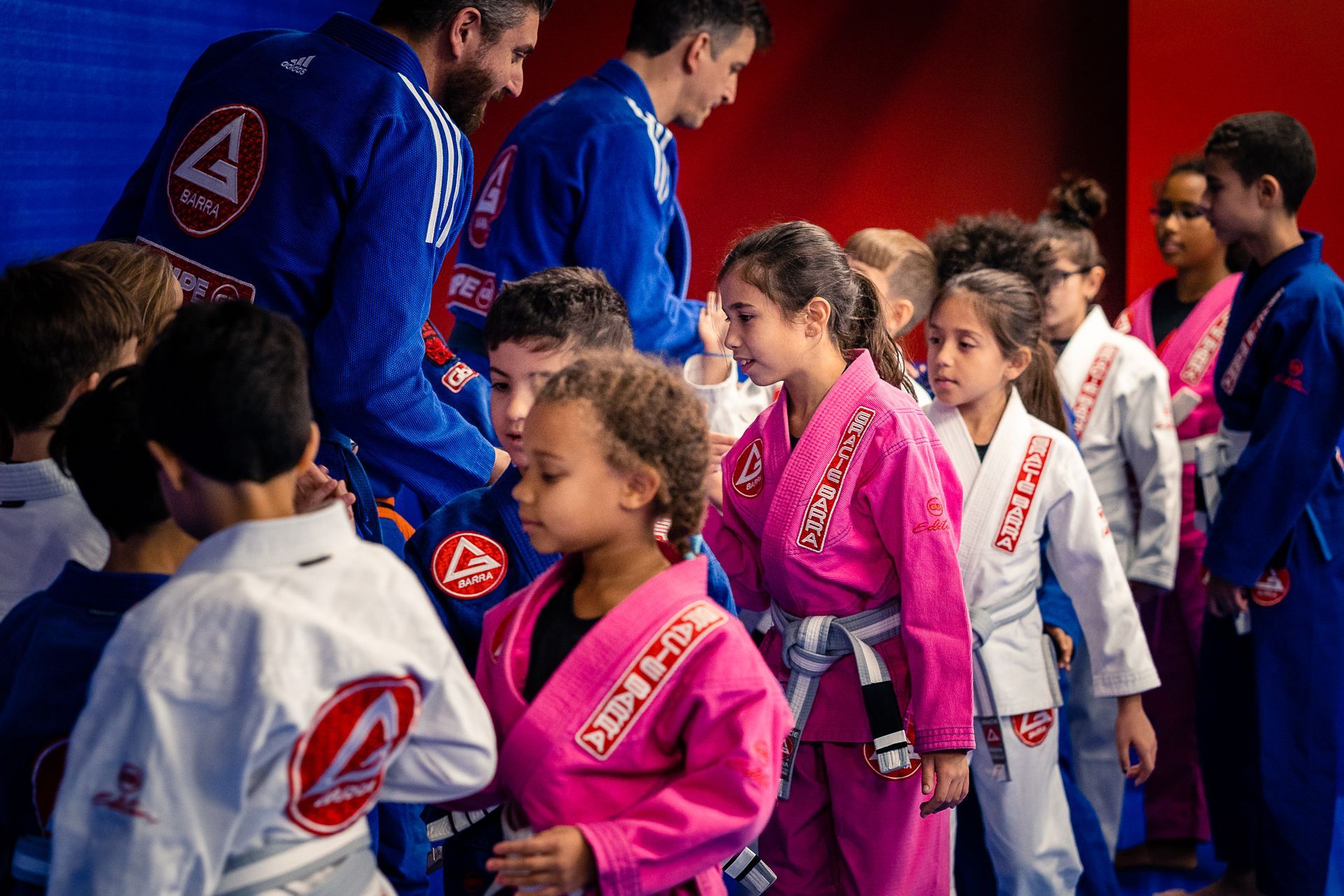Kids in pink and white martial arts uniforms line up for a ceremony, two men in blue uniforms in the background.