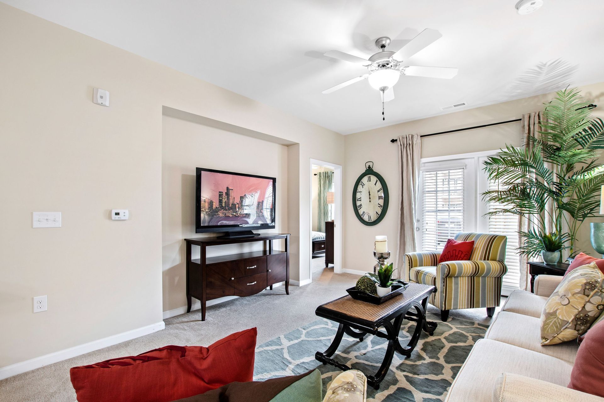 Living room in an apartment with beige walls, ceiling fan, TV, sofa, striped chair, and a tall plant.