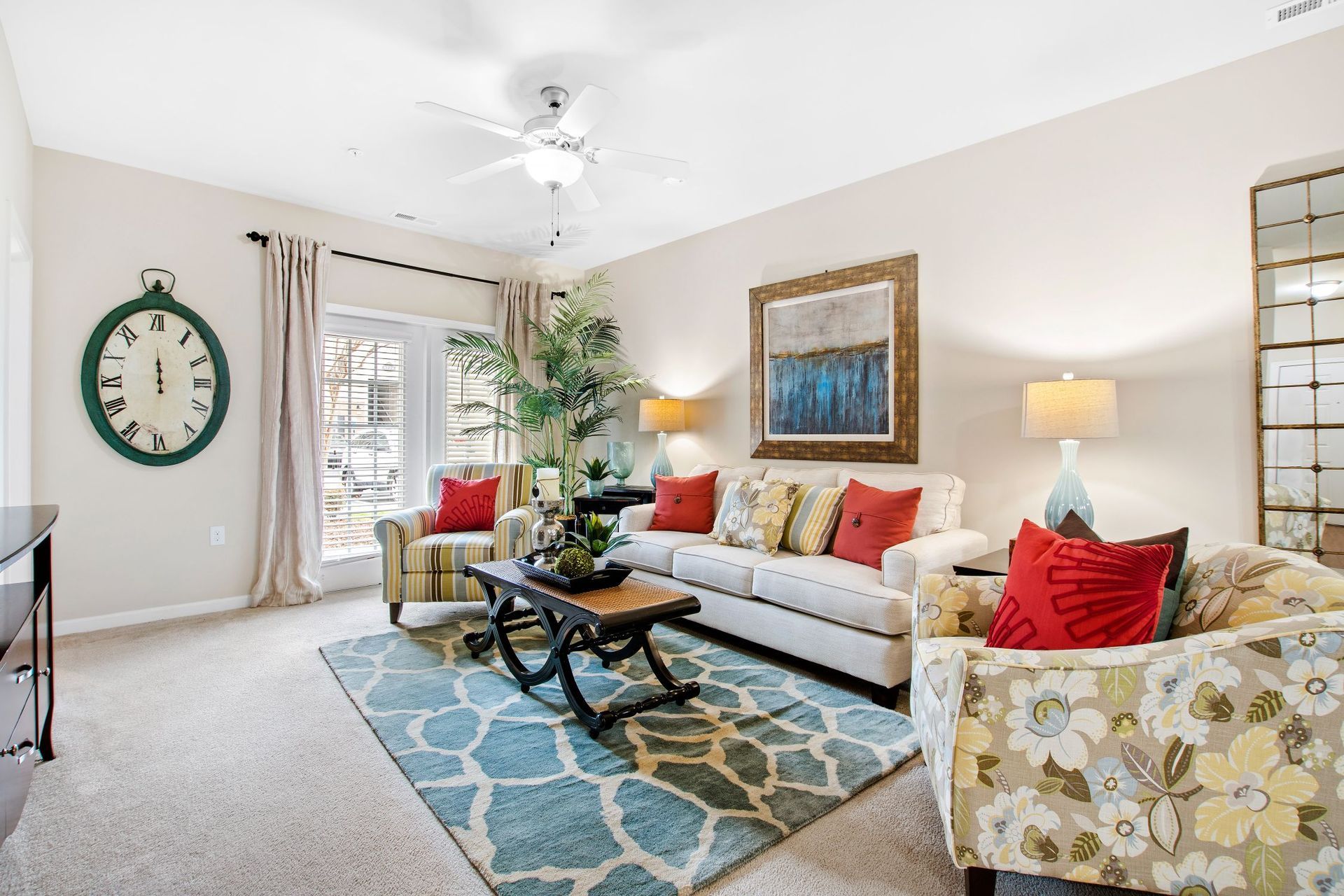 Bright living room with beige walls, ceiling fan, sofa, armchairs, plant, large clock, and blue patterned rug.