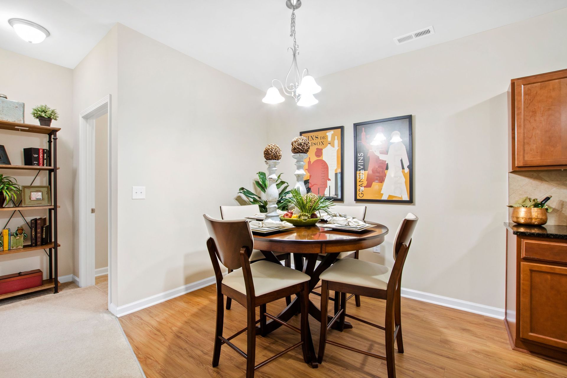 Dining area with round wooden table and four chairs, wall art, and a bookshelf to the left.