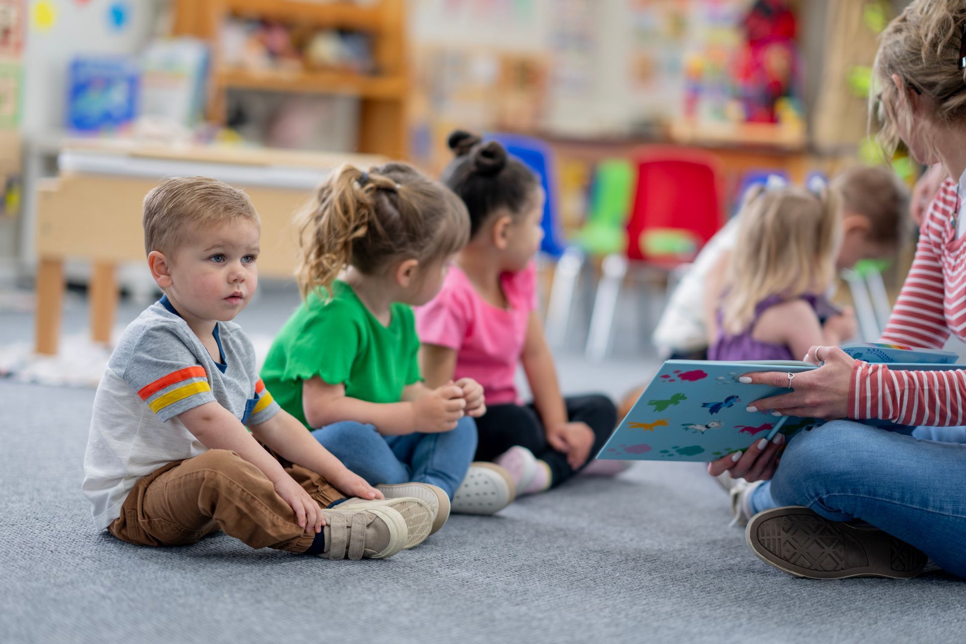 A group of children gathered in a preschool setting, listening to a story read by a teacher.