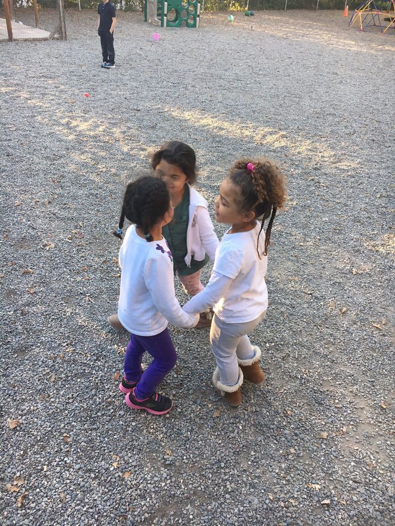 Three little girls are holding hands in a circle in a gravel playground.