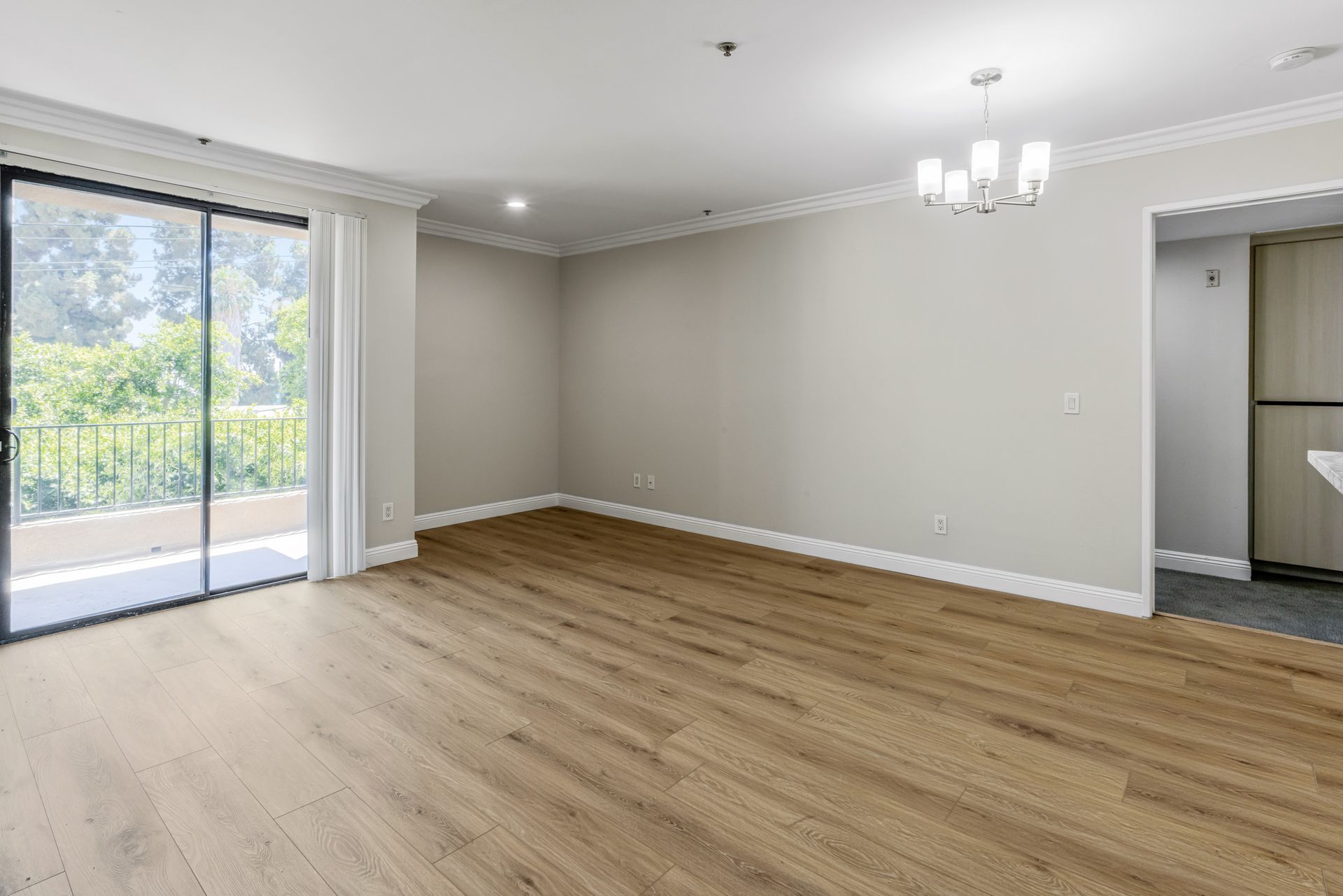 Empty living room with wood flooring, sliding glass door to balcony, neutral walls, and chandelier.