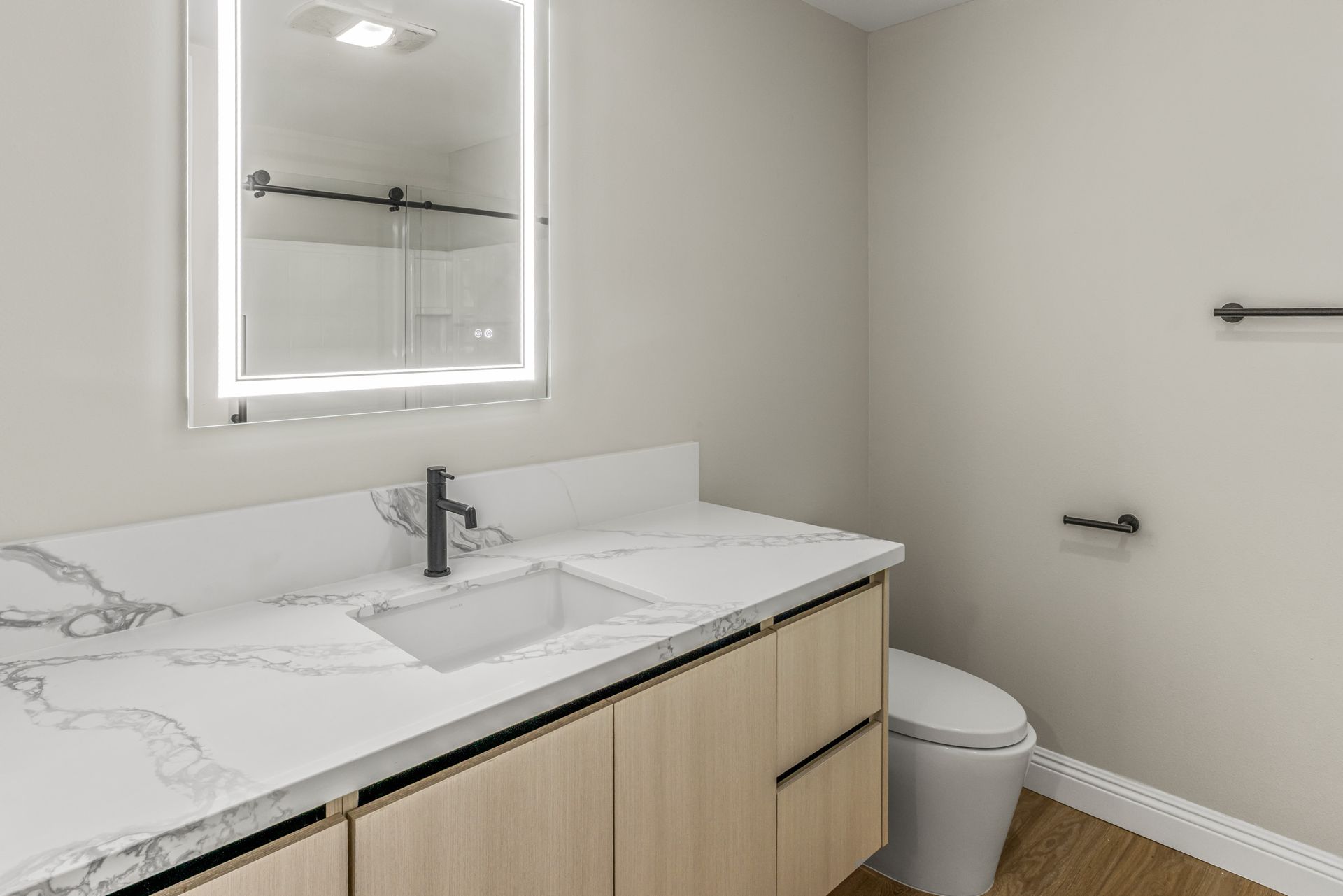 Modern bathroom with light wood vanity, white countertop, black fixtures, and a backlit mirror.
