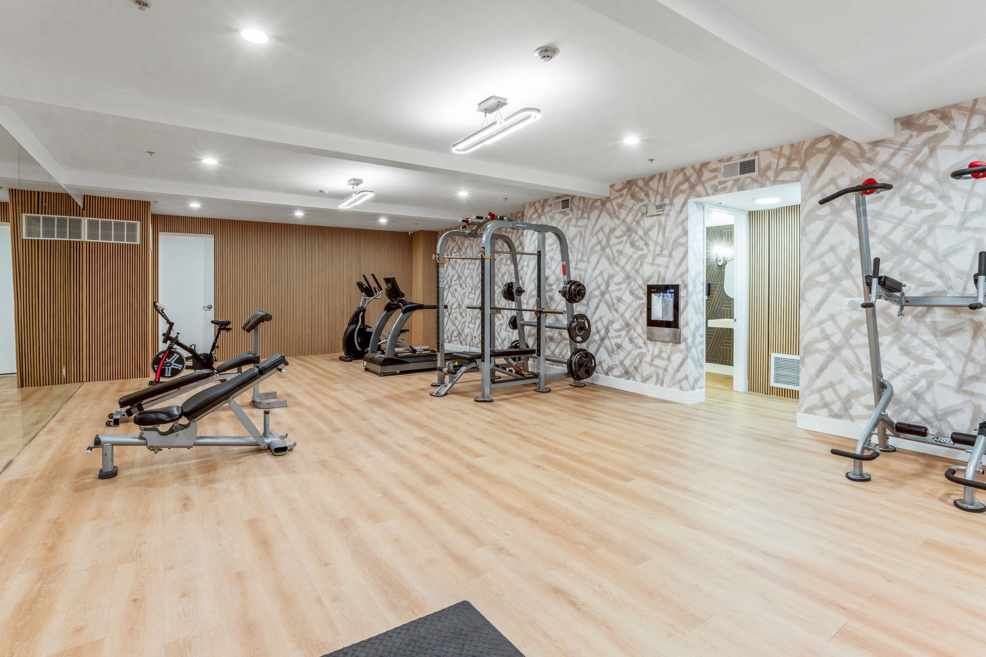Gym with exercise equipment on wood floor; weights, treadmill, stationary bikes.
