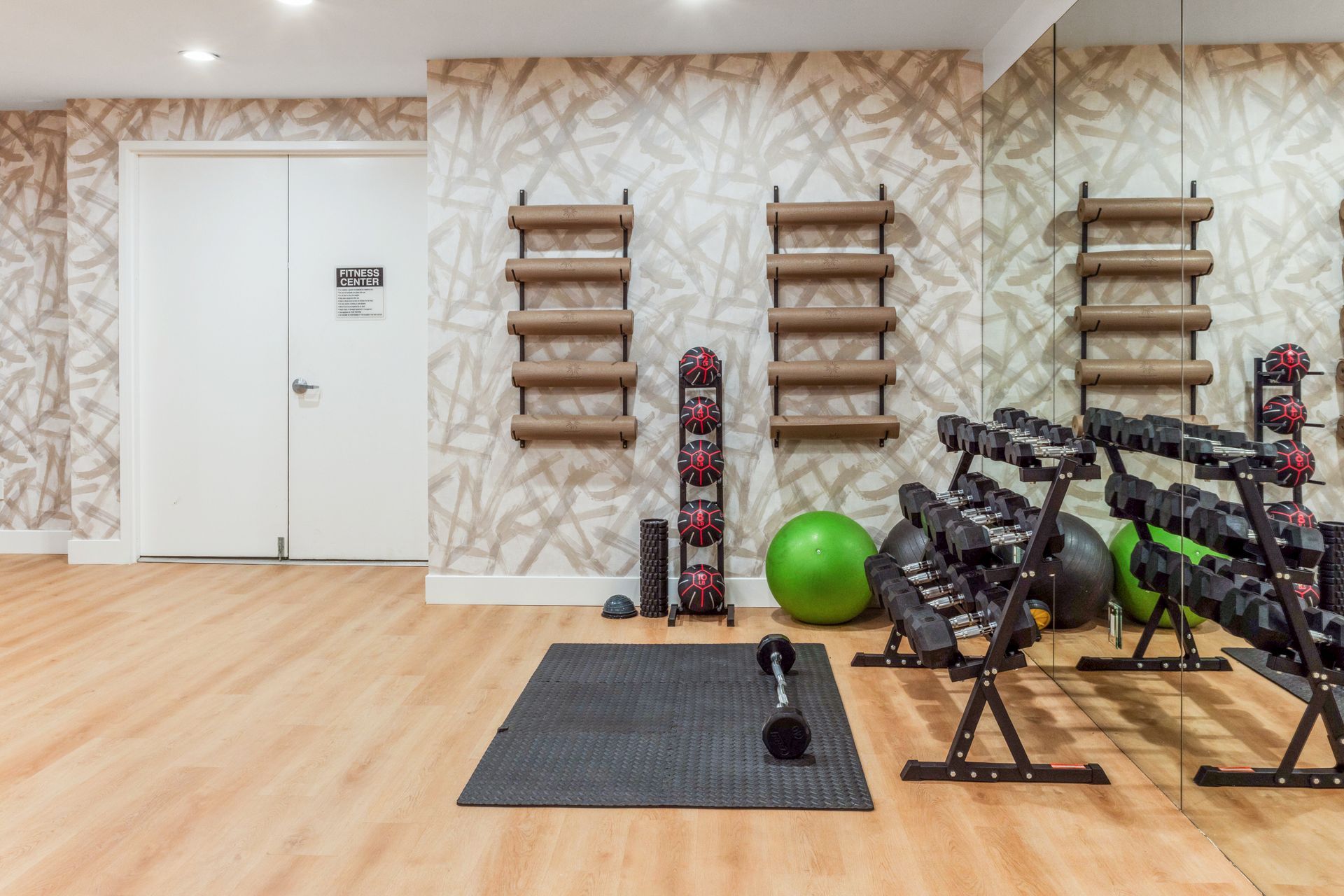 Gym with dumbbells, yoga balls, and mats on wood floor, with door and mirror.