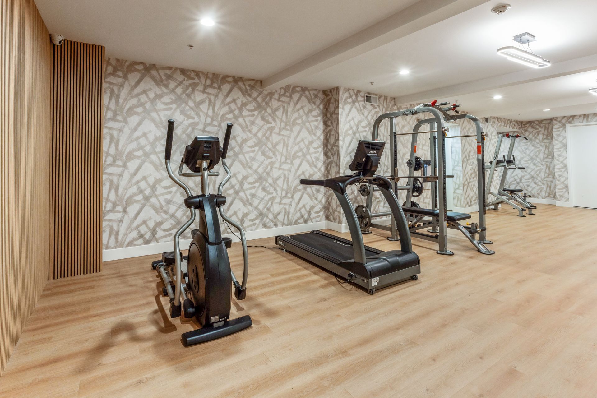 Gym with elliptical, treadmill, and weight machine on light wood floors, beige patterned wall.