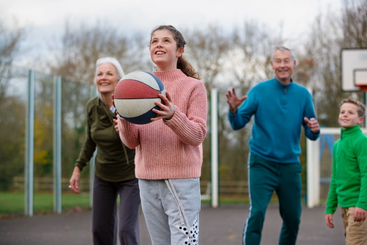Family playing basketball outdoors. Teen holding ball, others smiling, court and trees in background.