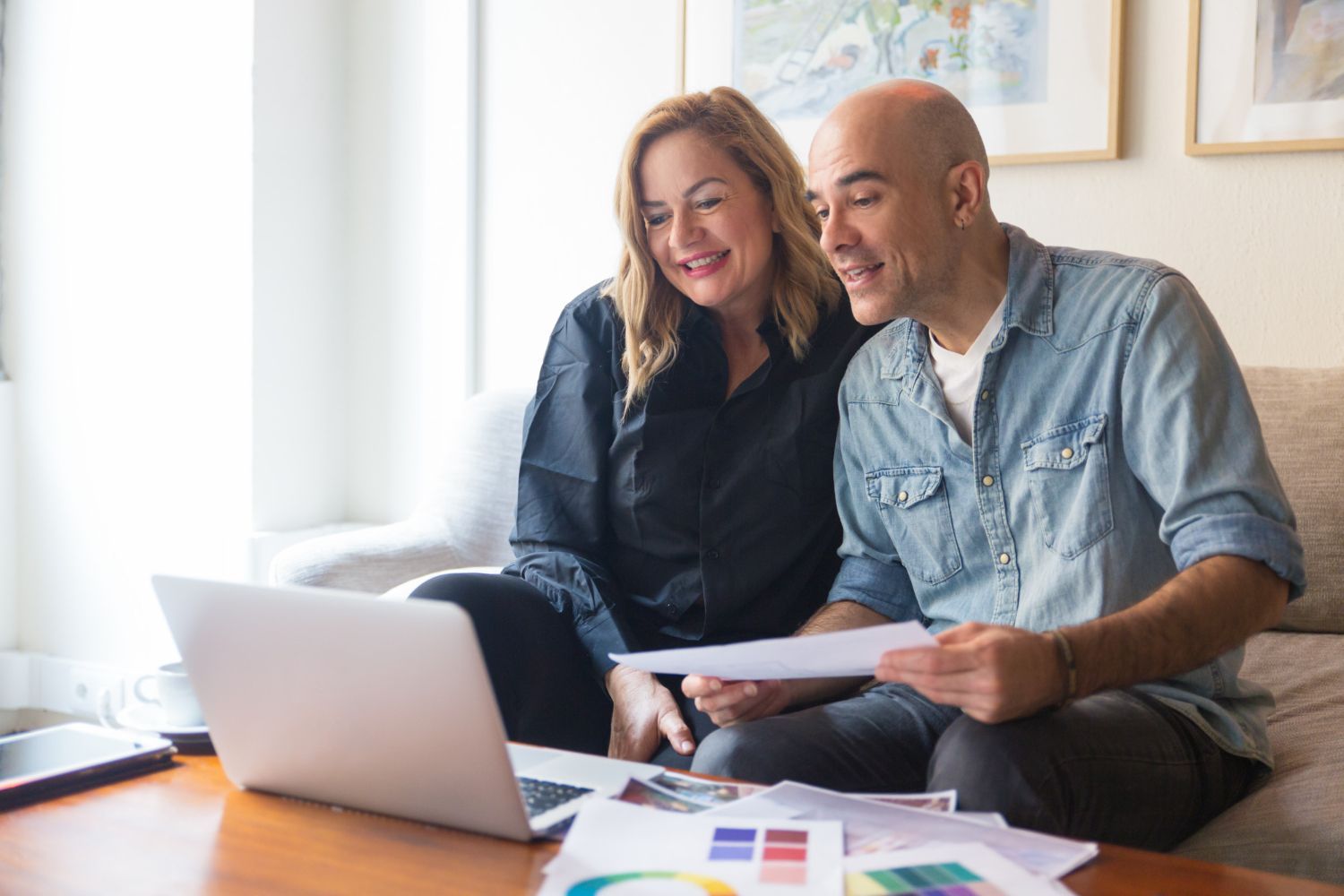 Couple on sofa looking at laptop, reviewing papers, smiling. Bright living room.