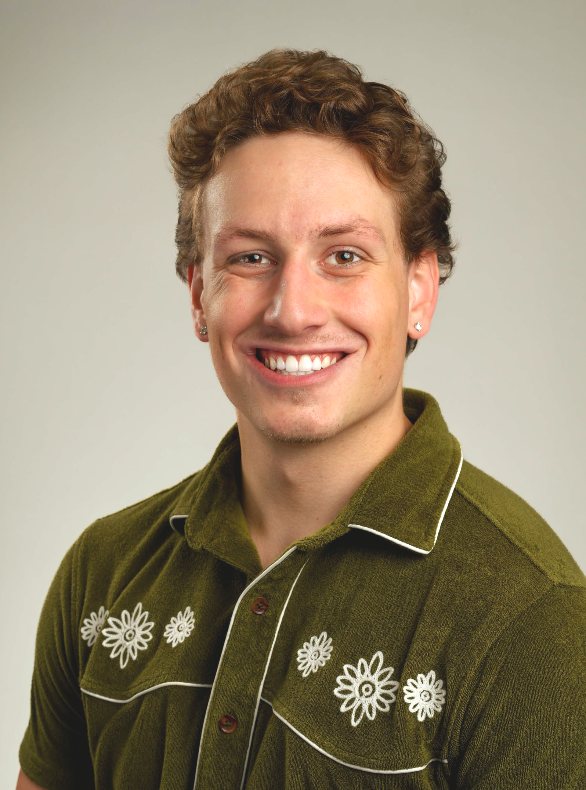 Man with curly brown hair smiles, wearing a green shirt with white floral embroidery.