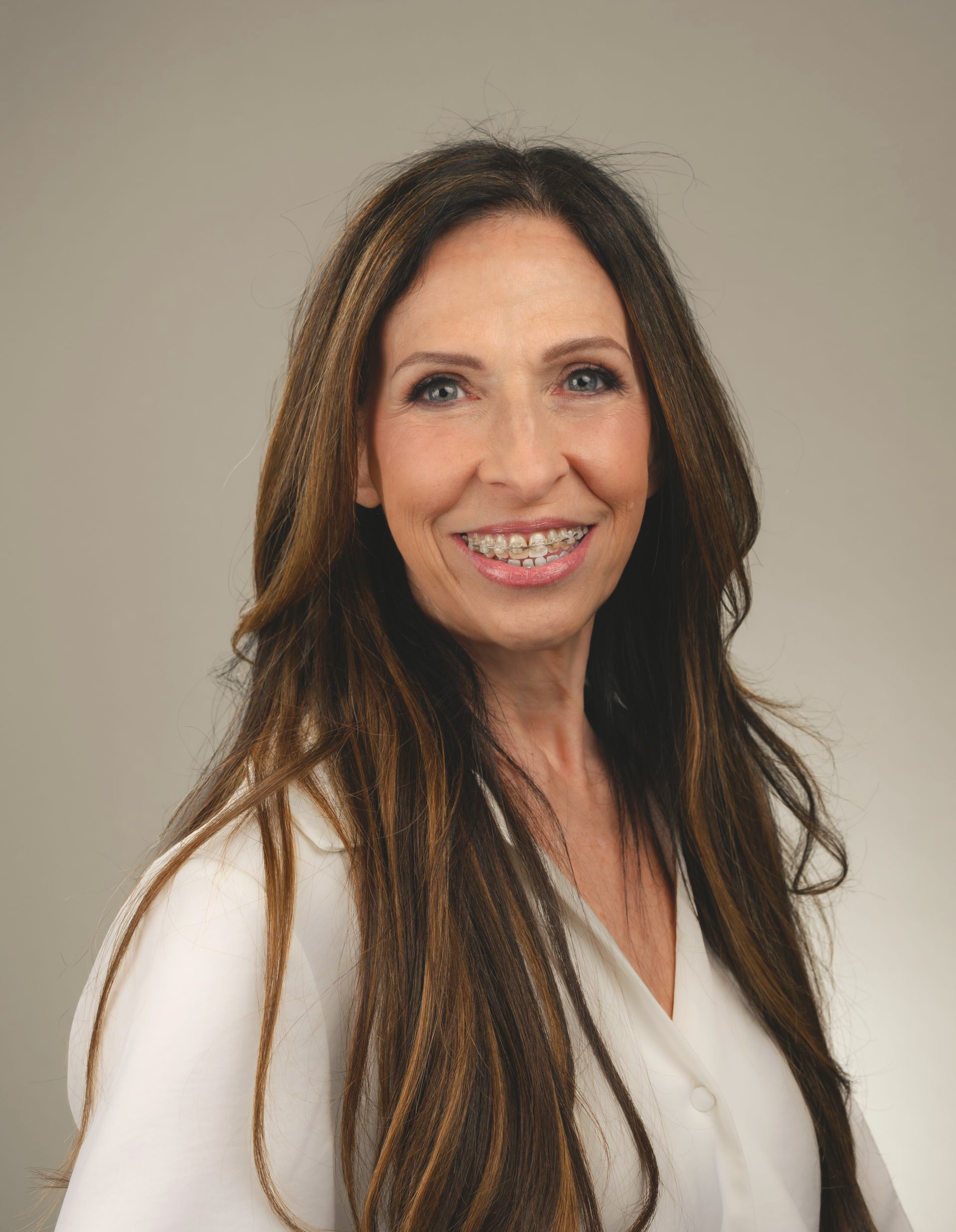 Woman with long brown hair smiling, wearing a white top, against a neutral background.