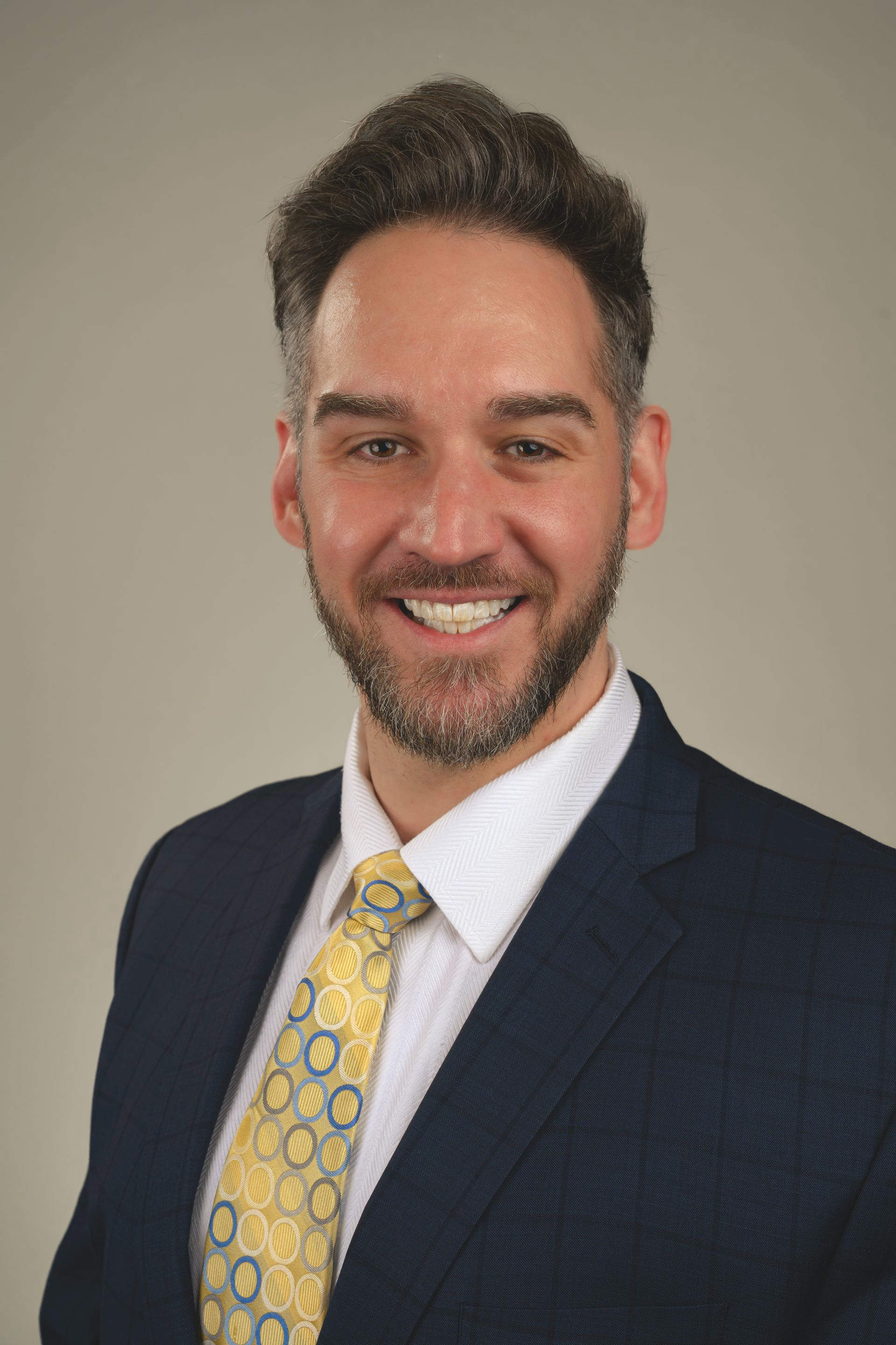 Man in a blue blazer, white shirt, and yellow patterned tie smiles. Gray background.