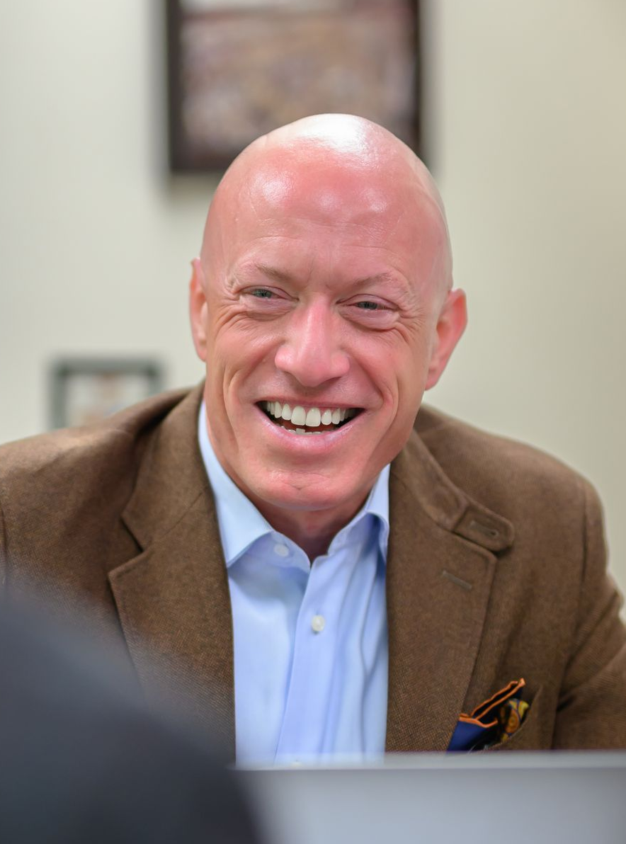 Bald man smiling, wearing a brown blazer over a light blue shirt. Indoor setting.