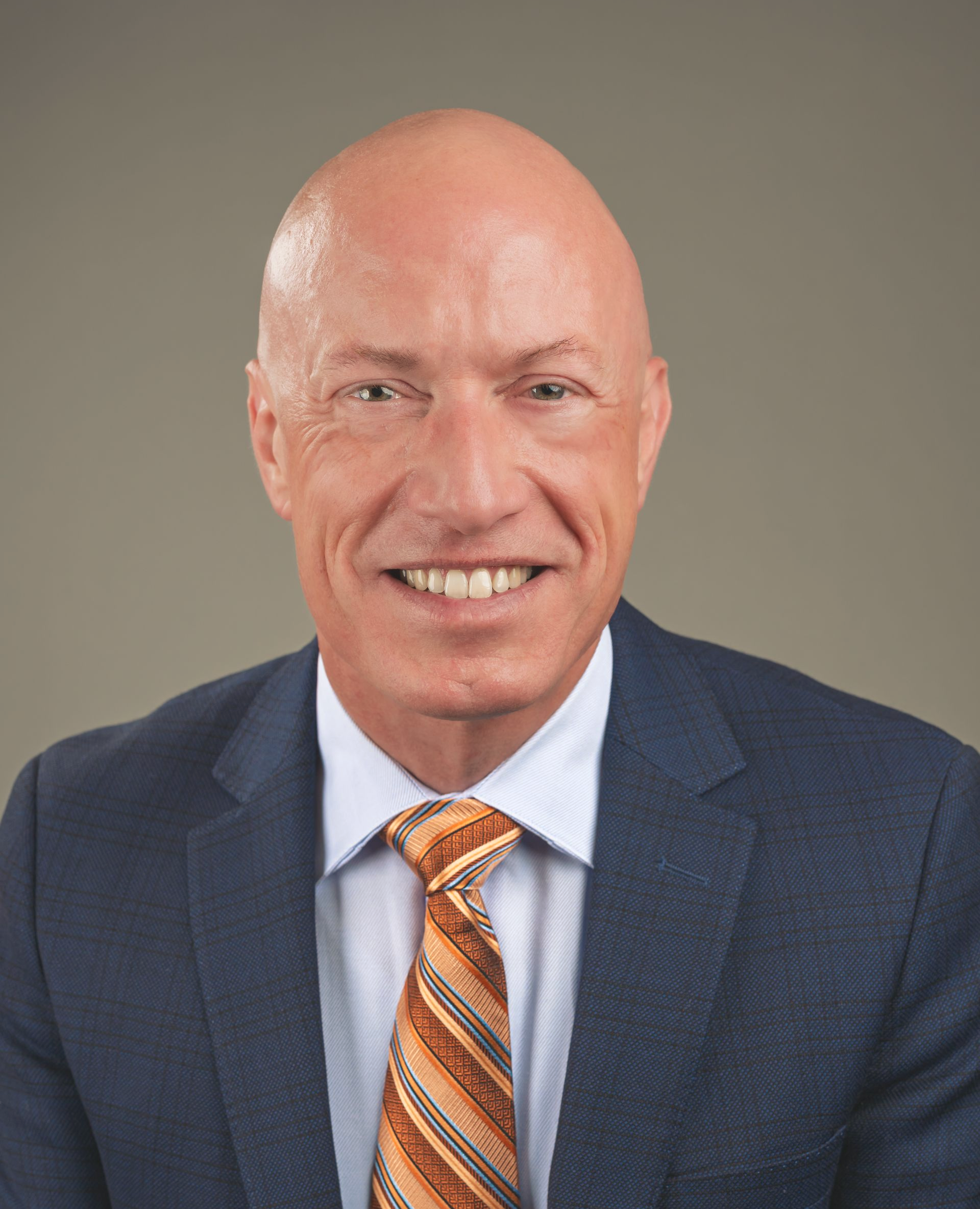 Man in a blue blazer and patterned tie smiles, against a gray backdrop.