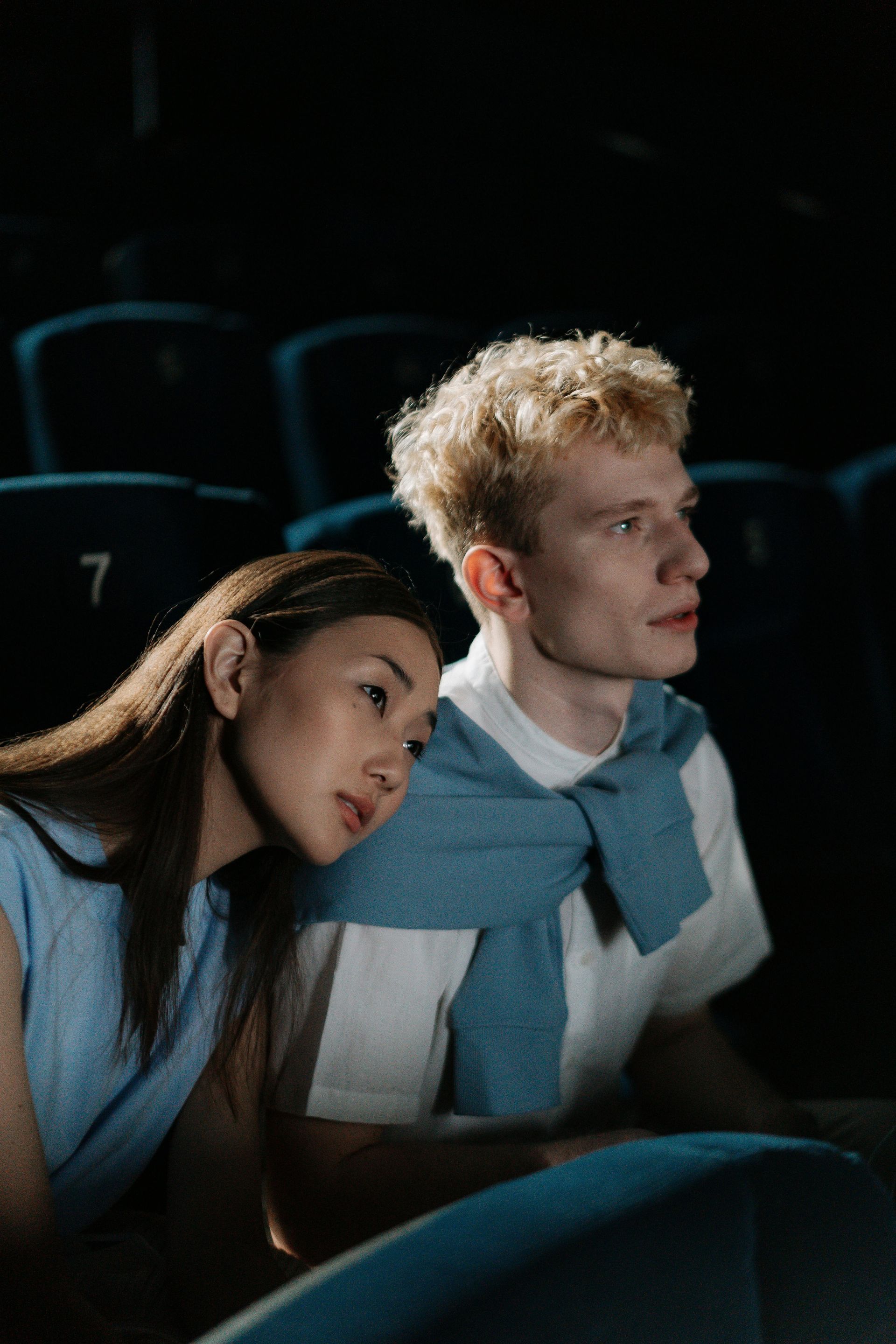 Two people seated in a dark theater, one resting their head on the other's shoulder, both wearing blue sweaters.