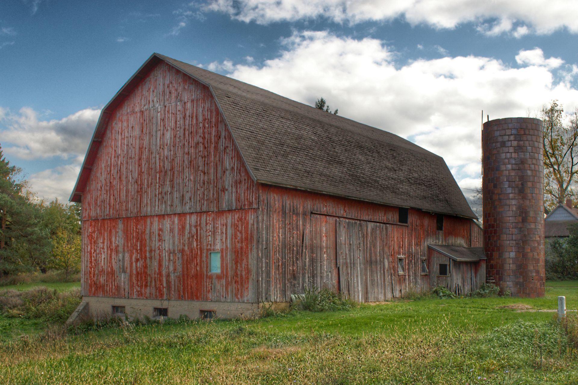 A red barn with a silo in the background is sitting in the middle of a grassy field.