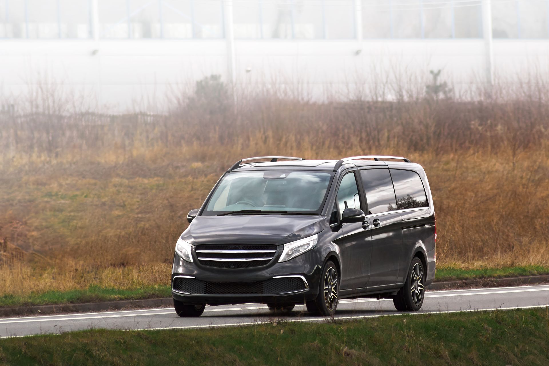 Une minifourgonnette noire circule sur une route mouillée, bordant un champ herbeux par temps brumeux.