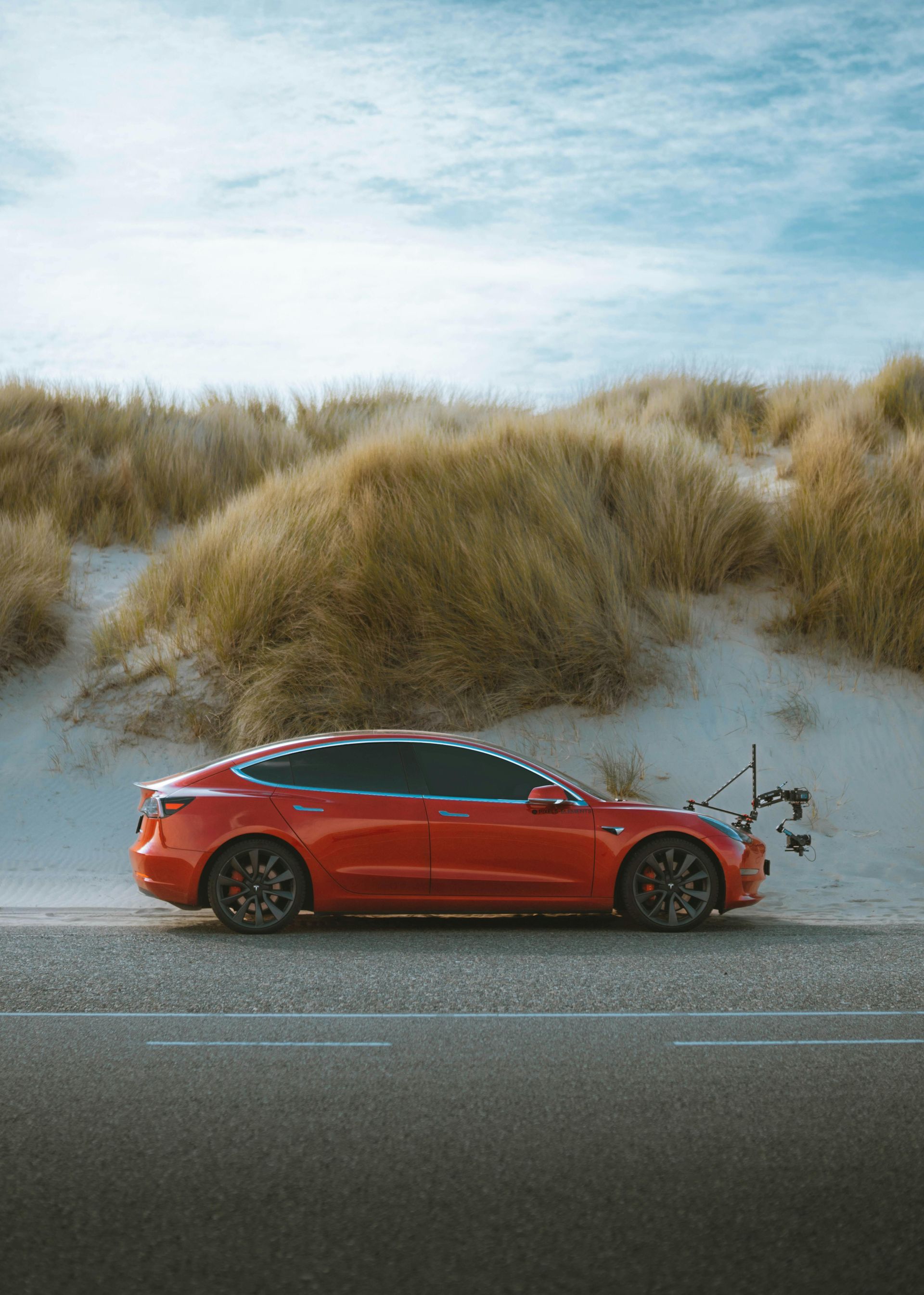 Voiture rouge roulant sur une route bordée de dunes de sable et d'herbe sous un ciel nuageux