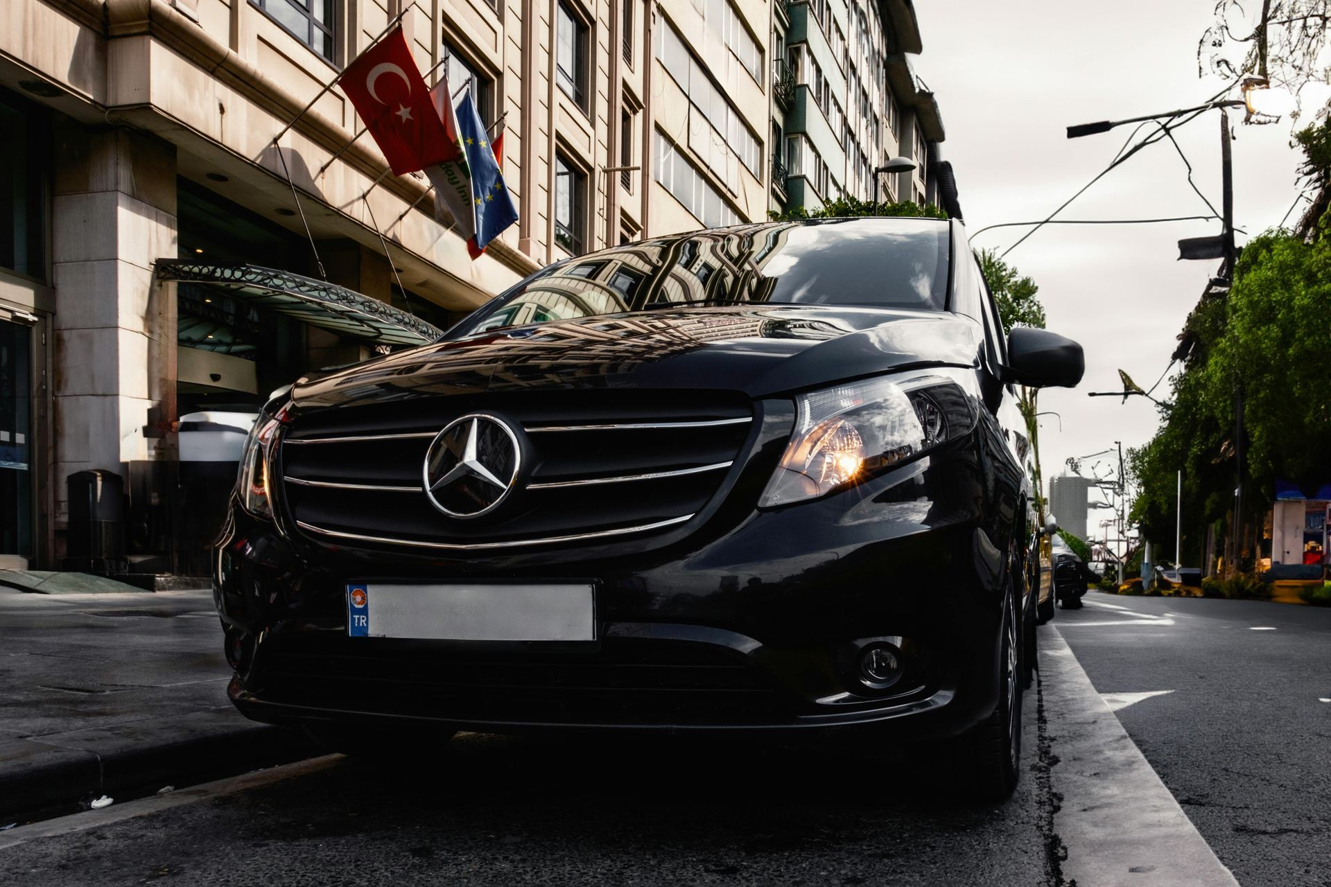 Une Mercedes-Benz noire est garée dans une rue de la ville, à côté de bâtiments et de drapeaux.