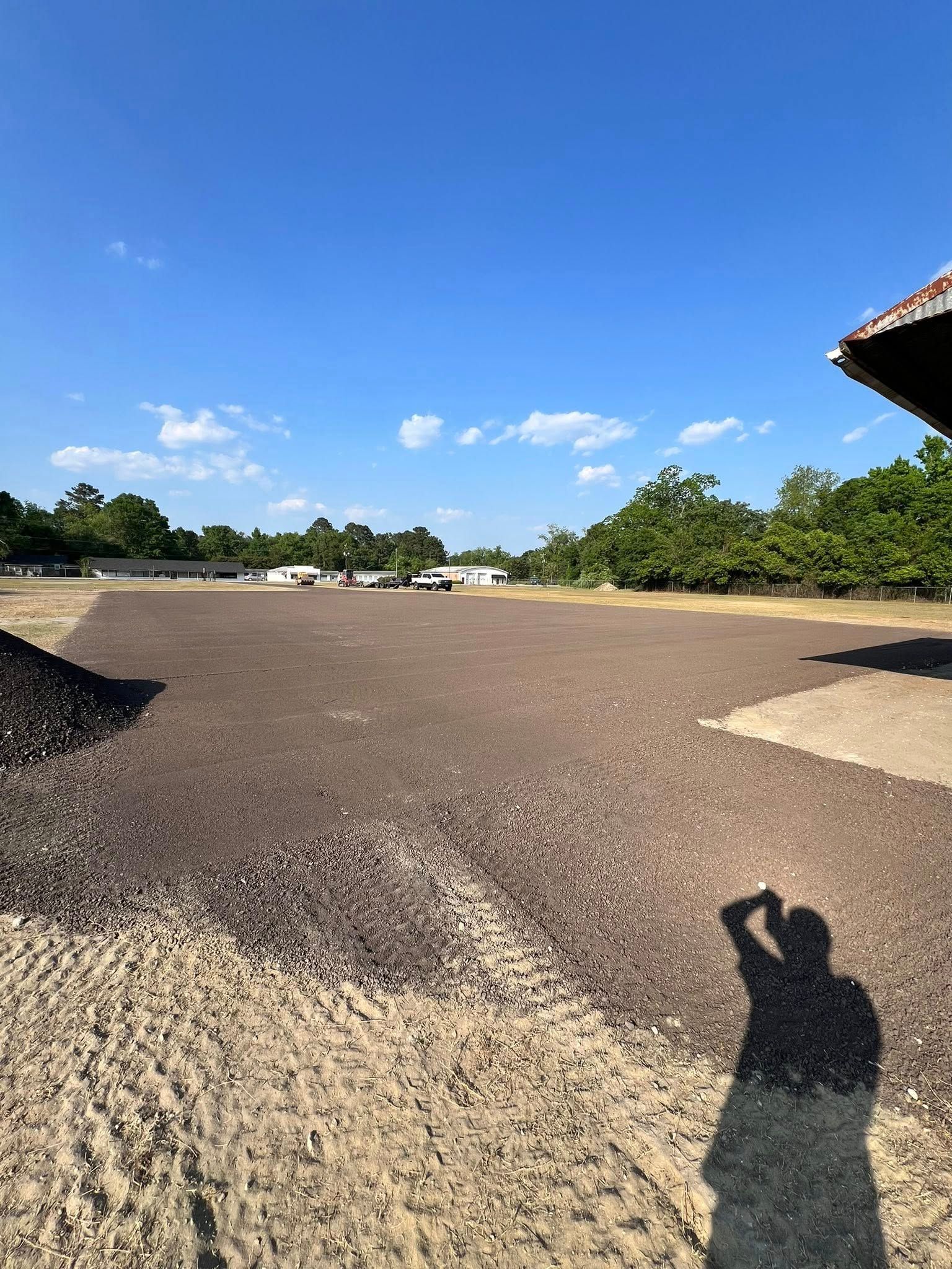 Wide gravel lot under a bright blue sky with a few clouds and tree-lined edges, shadow in foreground