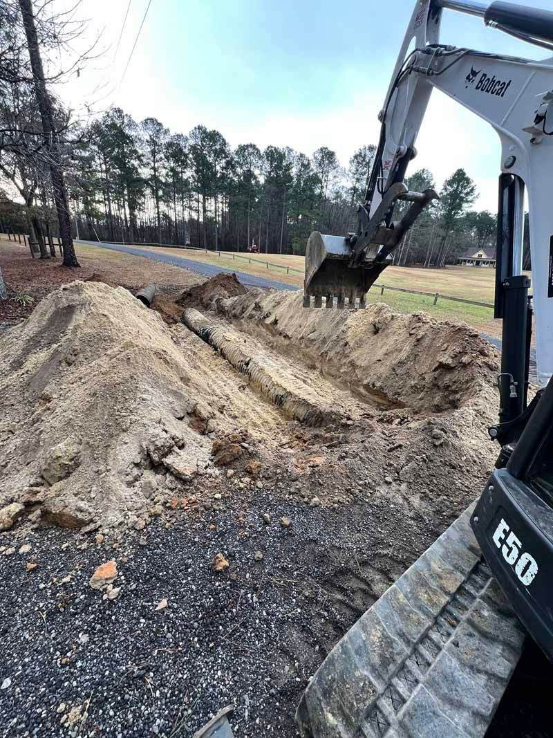 Excavator digging a muddy trench beside a wooded construction site