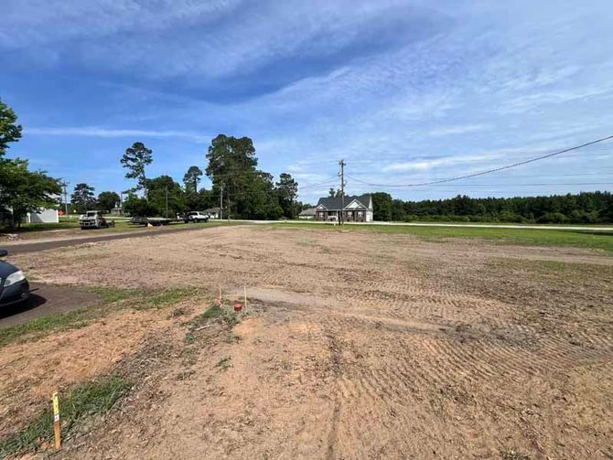 Cleared dirt lot under blue sky beside a road, with trees and utility lines in the distance.