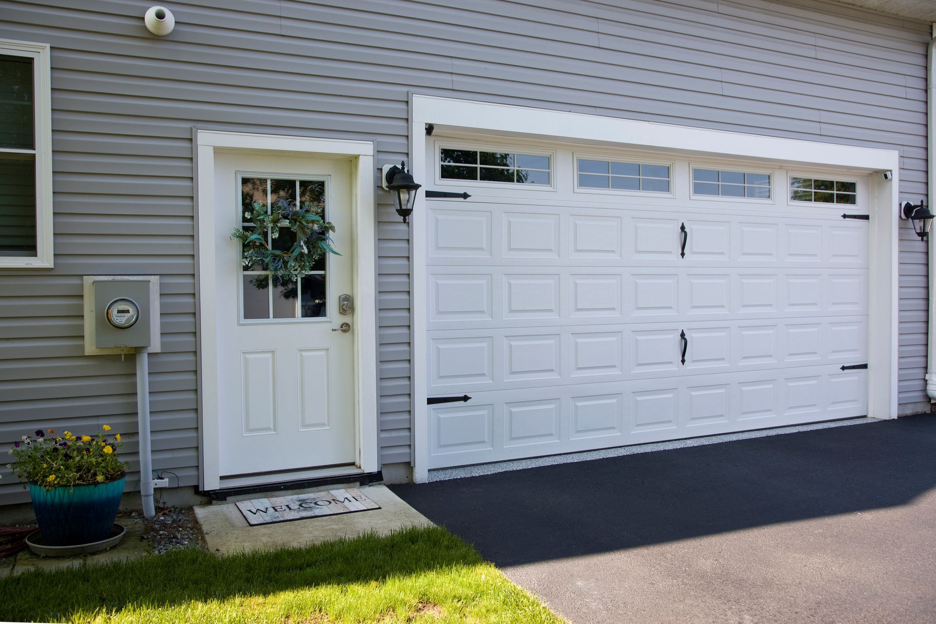 White garage door and matching door next to light blue siding. Potted flowers sit to the left. White garage door and matching door next to light blue siding. Potted flowers sit to the left.