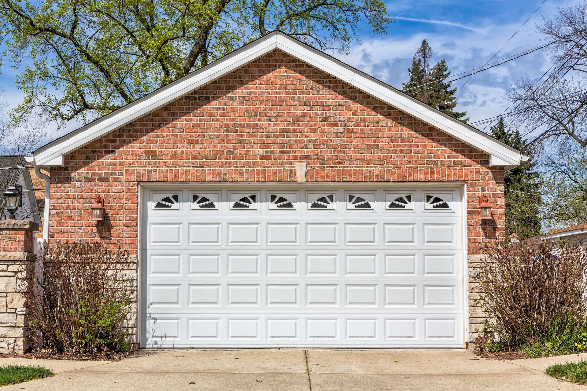 Brick garage with white garage door and decorative top panels, set against greenery and blue sky.