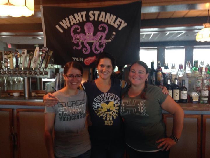 Three women pose in front of a sign that says i want stanley
