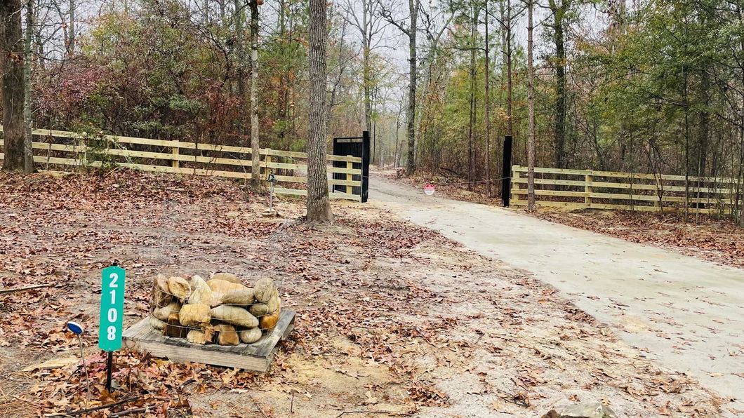 A pile of logs is sitting on the side of a dirt road next to a wooden fence.