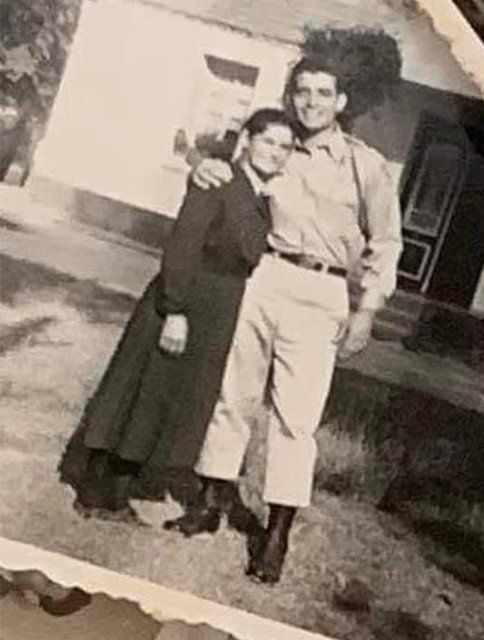 Couple standing outside a building; man has arm around woman. They are dressed in light-colored pants and coat.