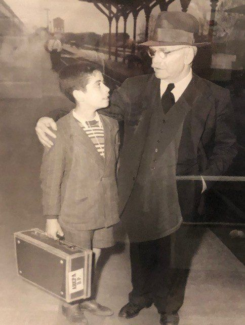 Boy with suitcase looks at man in a suit, standing on a train platform.
