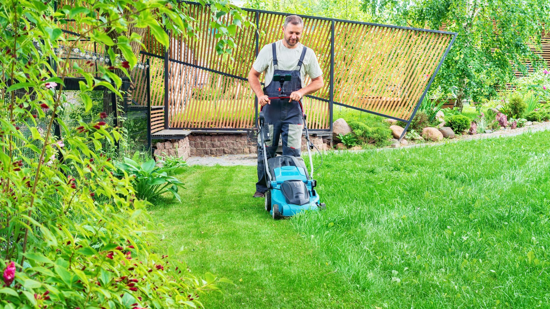 Man in overalls mowing a green lawn with a teal and black lawnmower. Lush garden in the background.