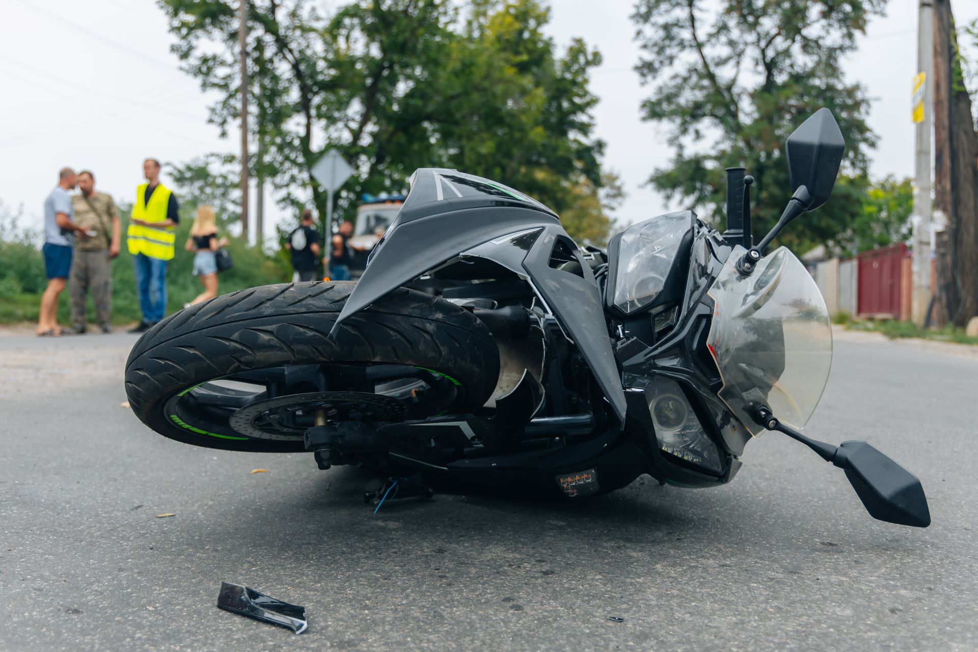Motorcycle crashed on a road. People gather in the background, near trees. Gray asphalt, dark bike.
